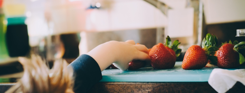 Ett barn sträcker sig upp med handen på en diskbänk. På diskbänken ligger skärbräda och jordgubbar.